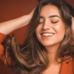 Gros plan d'une femme souriante, les yeux fermés, passant les mains dans ses longs cheveux châtains et doux