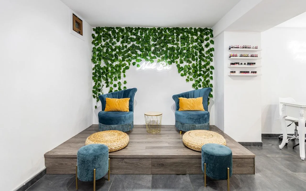 Un coin onglerie avec un mur de feuillage vert en cascade et une petite enseigne néon, deux fauteuils en velours bleu canard ornés de coussins moutarde sur une estrade en bois, des poufs repose-pieds tressés, et des étagères de vernis colorés sur la droite.