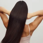 Vue de dos d’une femme en haut blanc à fines bretelles, les bras levés, passant les doigts dans ses longs cheveux bruns brillants
