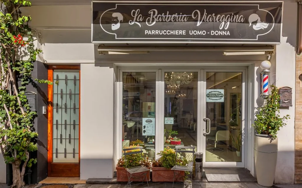 La devanture de La Barberia Viareggina par une journée ensoleillée, avec une enseigne de barbier classique rouge, blanche et bleue à droite des portes vitrées. Des plantes en pot et une végétation grimpante encadrent l'entrée, et un lustre est visible à l'intérieur à travers les vitres.