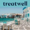 Le front de mer de la Petite Venise à Mykonos : maisons blanchies à la chaux aux balcons de bois colorés au bord de l'eau, la mer Égée turquoise au loin, et au premier plan des tables de café blanches et bleu canard installées sur une terrasse pavée.