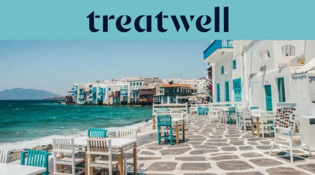 Le front de mer de la Petite Venise à Mykonos : maisons blanchies à la chaux aux balcons de bois colorés au bord de l'eau, la mer Égée turquoise au loin, et au premier plan des tables de café blanches et bleu canard installées sur une terrasse pavée.
