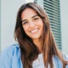 Une femme aux longs cheveux bruns et aux sourcils bien définis se tient à l'extérieur, souriante, devant un bâtiment aux volets verts