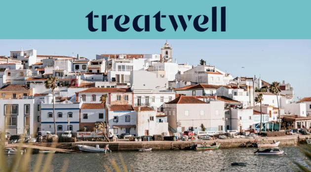 Un village de pêcheurs blanchi à la chaux de l'Algarve, aux maisons aux toits de terre cuite s'étageant à flanc de colline au-dessus d'un plan d'eau calme, avec de petits bateaux de pêche amarrés le long du quai, des palmiers et le clocher d'une église sous un ciel bleu limpide.
