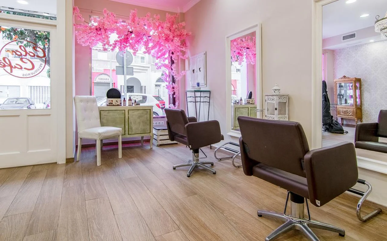 L'intérieur de Color Café Peluqueros à Torremolinos, un salon de coiffure aux murs roses avec des fauteuils marron, de grands miroirs et une composition de cerisier en fleurs rose en vitrine.