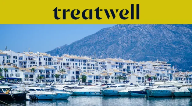 Le port de plaisance de Puerto Banús, à Marbella, bordé d'immeubles blancs et de yachts amarrés sur fond de montagnes, sous une banderole jaune Treatwell.