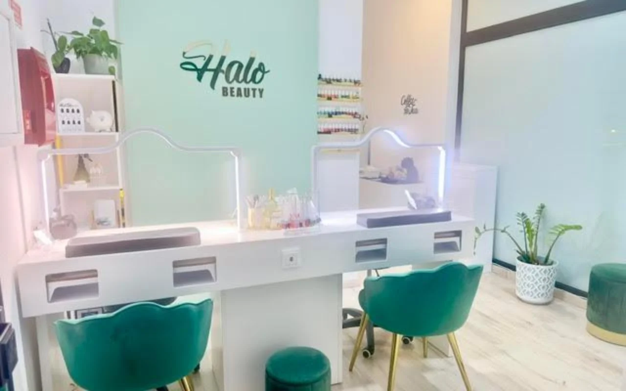 Un salon d'ongles avec un mur d'accent vert menthe portant le logo Halo Beauty, un double plan de manucure blanc avec des séparations en perspex éclairées, deux fauteuils tonneau en velours vert émeraude, un présentoir mural de vernis à ongles et des plantes en pot.