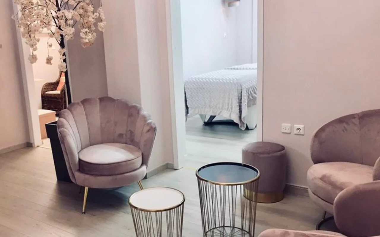 L'espace d'attente rose poudré de Nomikou Antonia Aesthetic Lab à Syros, avec un fauteuil en velours à dossier festonné, de fines tables d'appoint dorées, une branche de cerisier en fleurs et une cabine de soin au-delà de la porte.