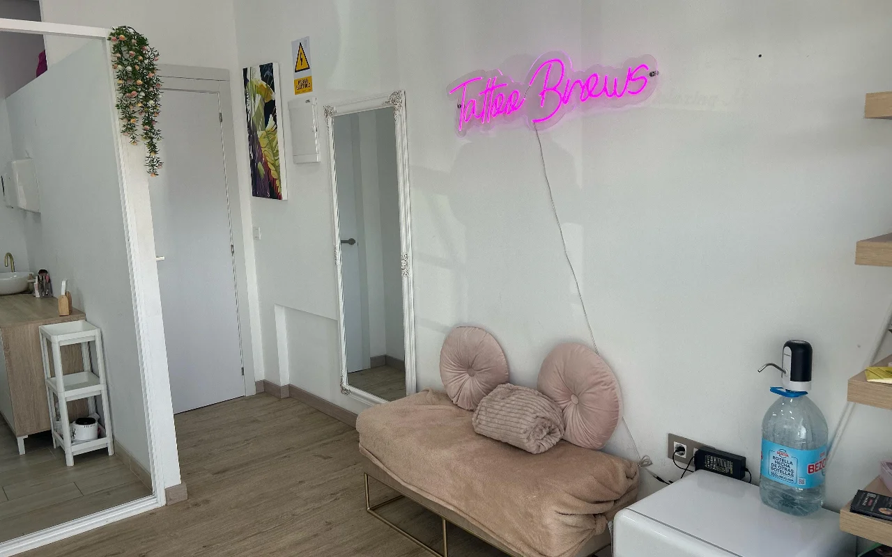 Un espace de soin chez YG Beauty Estudio à Torremolinos, avec une enseigne néon rose « Tattoo Brows », un miroir en pied et une méridienne beige capitonnée.