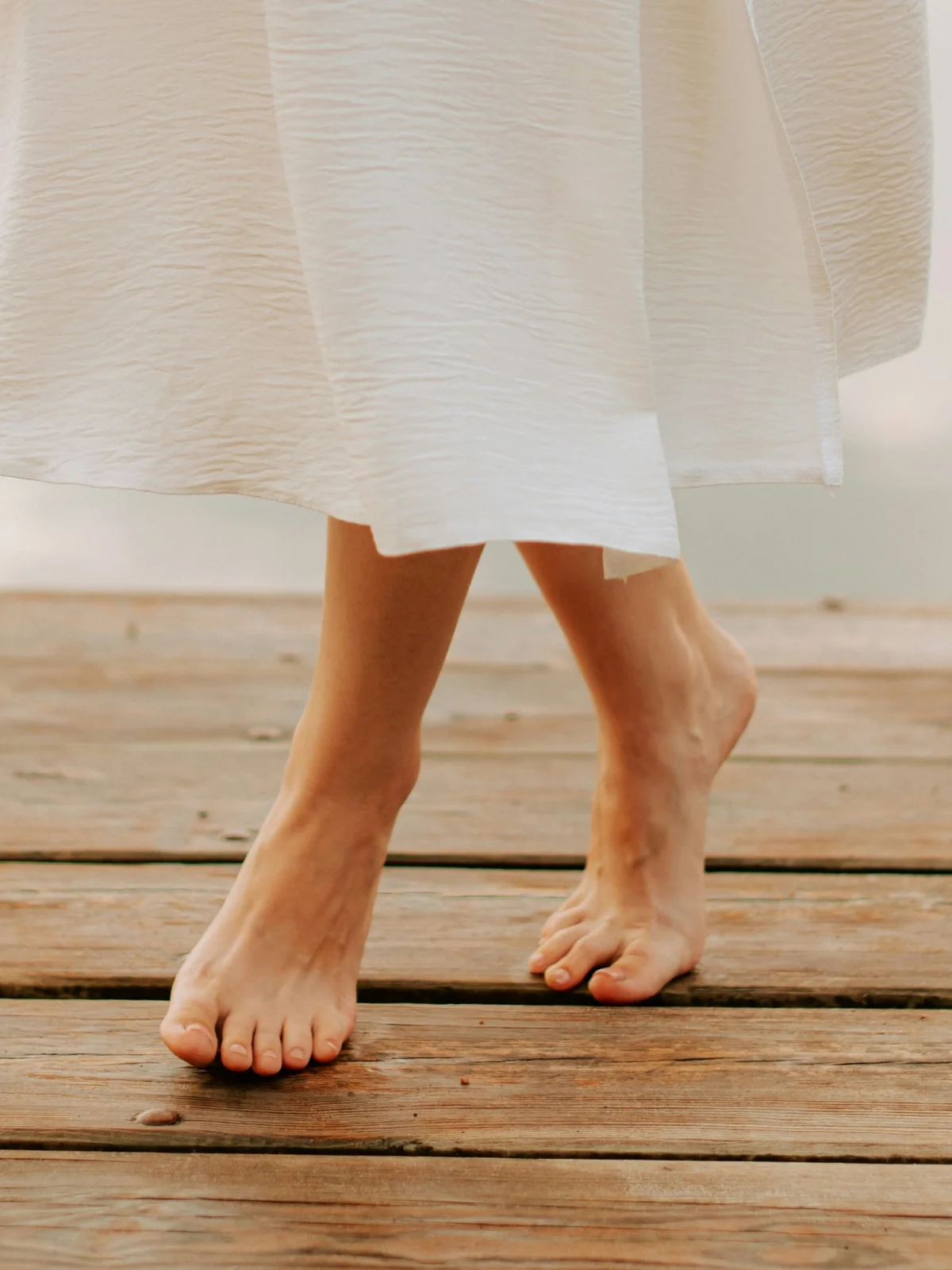 Gros plan sur les jambes d'une femme aux pieds nus et sains, vêtue d'une jupe blanche fluide, qui avance sur un ponton en bois au bord d'un lac