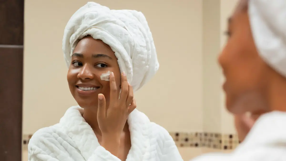 Une femme en peignoir blanc, une serviette blanche enroulée autour de la tête, sourit dans le miroir de la salle de bain en appliquant un SPF sur son visage