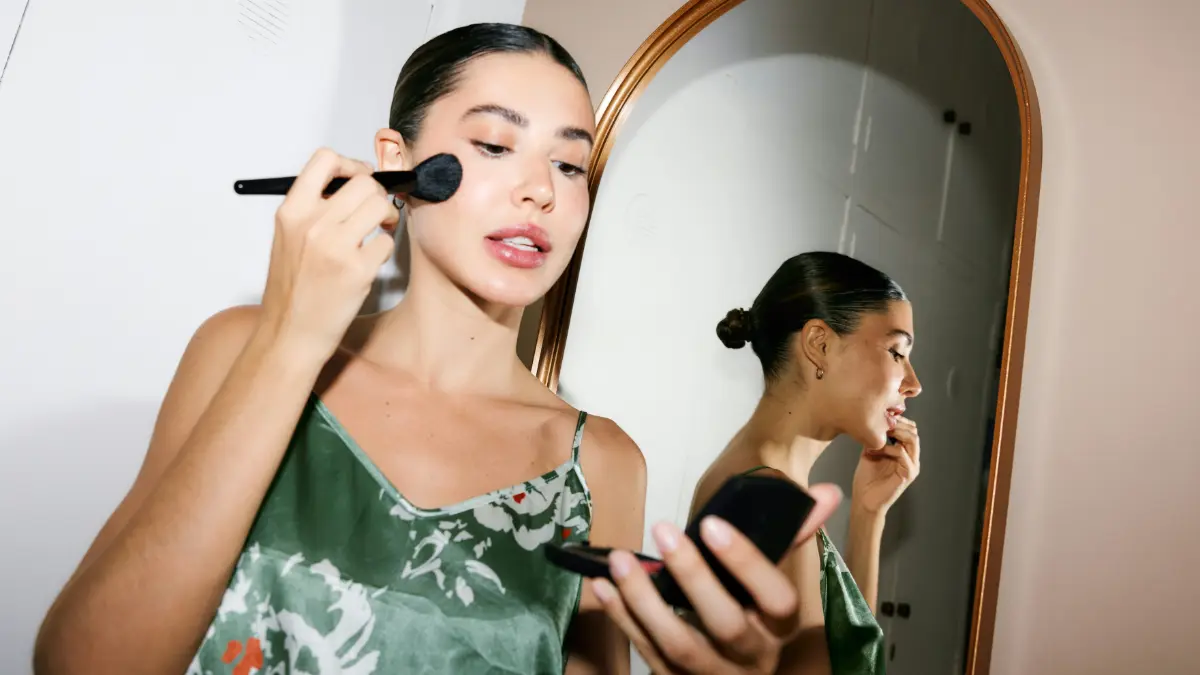 Une femme élégante prête à sortir se regarde dans un miroir de poche en appliquant son maquillage au pinceau