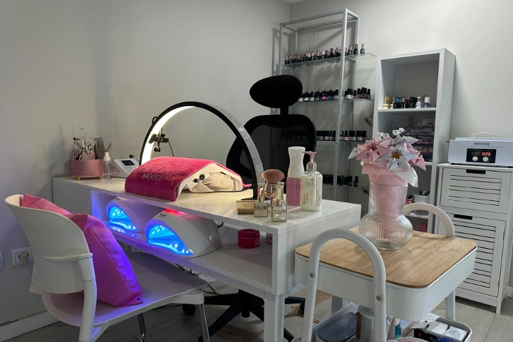 Poste unique de manucure avec un bureau blanc, une lampe ronde et deux lampes LED bleutées, une chaise noire et des étagères garnies de vernis colorés, égayé par un vase rose rempli de fleurs.