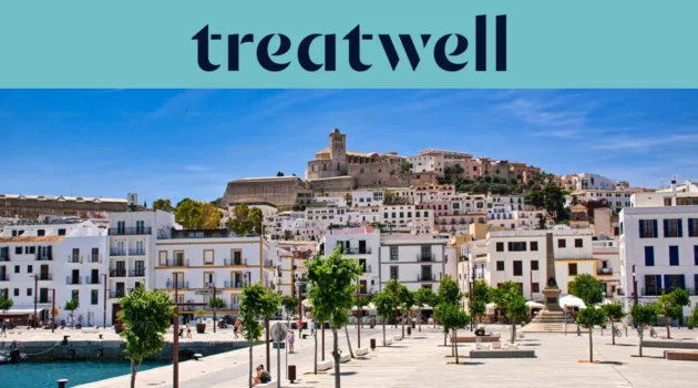 Vue ensoleillée sur Ibiza Ville, avec des bâtiments blanchis à la chaux qui grimpent jusqu'à la cathédrale et aux anciennes murailles de Dalt Vila sur la colline. Une promenade portuaire bordée d'arbres traverse le premier plan sous un ciel bleu limpide.