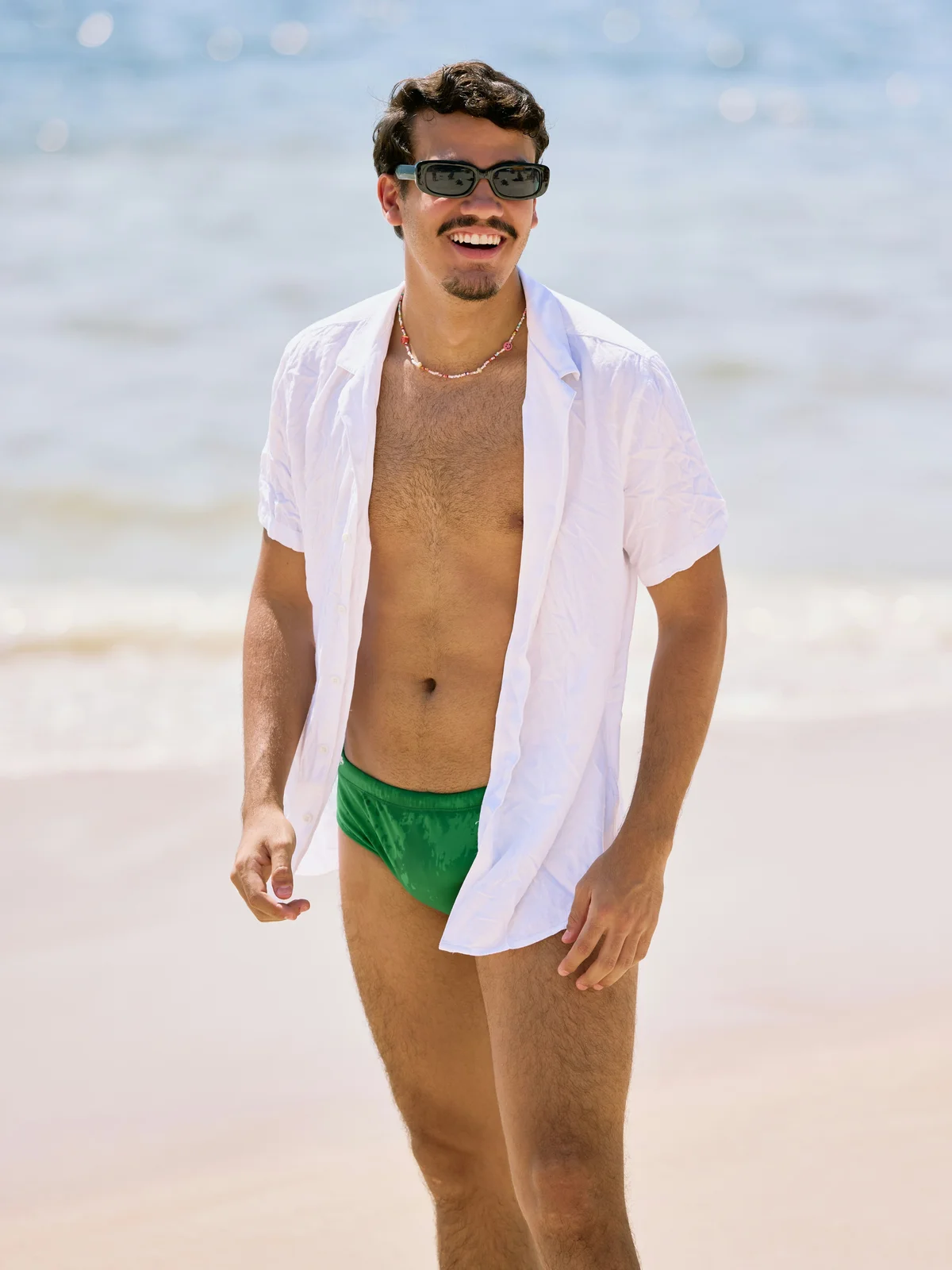 Un homme souriant se tient sur une plage par une journée ensoleillée ; il porte un maillot de bain vert, une chemise blanche ouverte et des lunettes de soleil noires