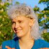 Een jonge vrouw met een krullend witblond mullet-kapsel zittend buiten op een zonnige dag.