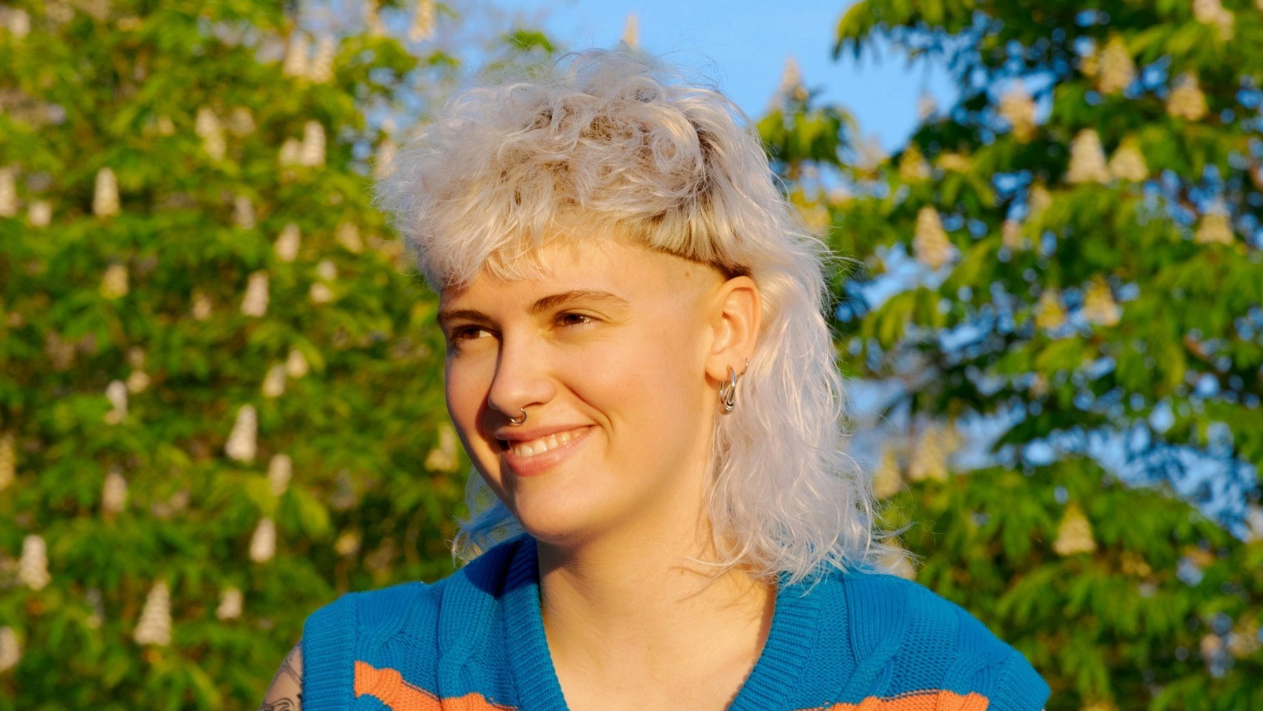 Een jonge vrouw met een krullend witblond mullet-kapsel zittend buiten op een zonnige dag.
