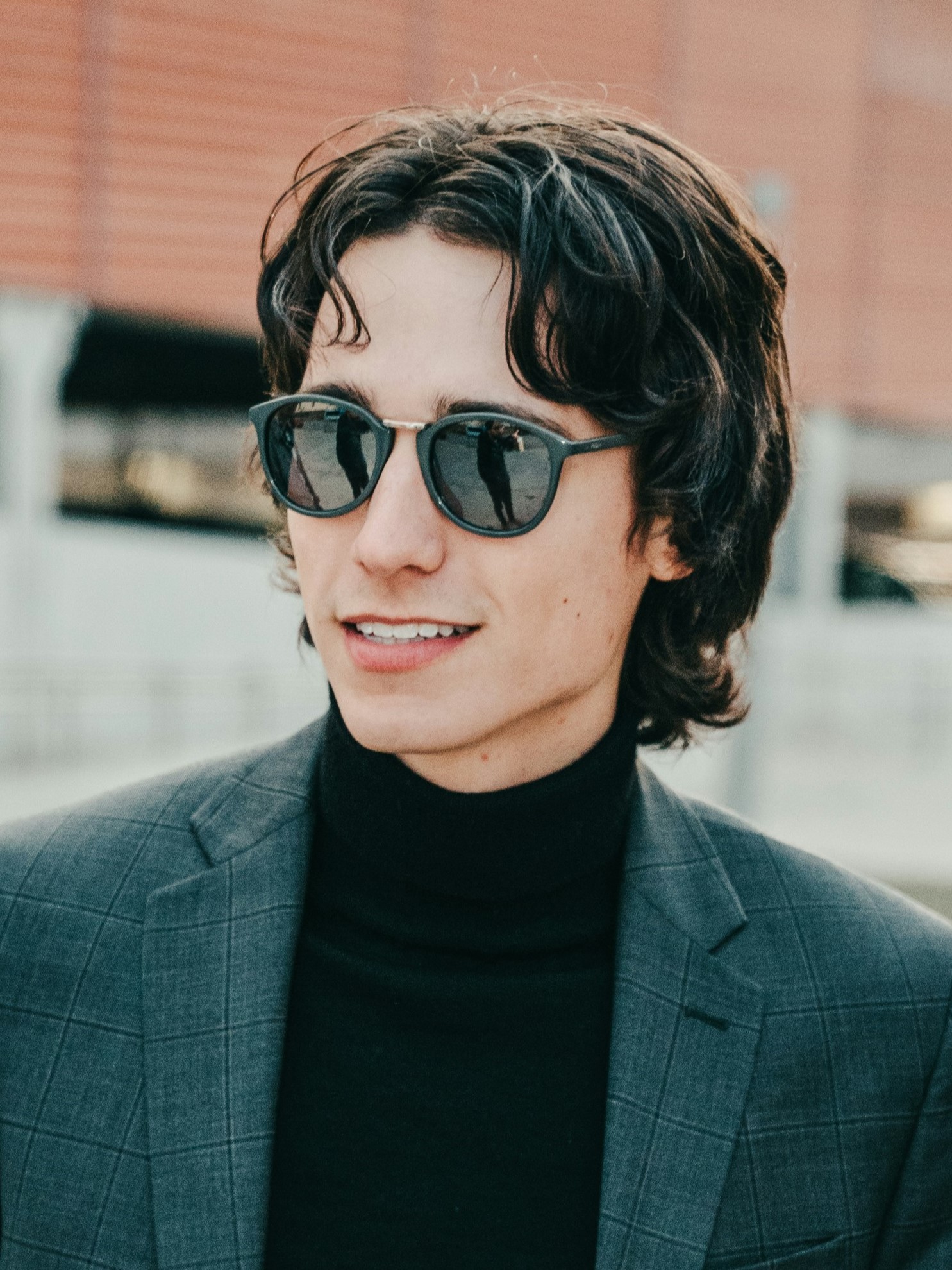 Een jonge man met een retro mullet-kapsel in een grijs colbert en zonnebril, lachend buiten.