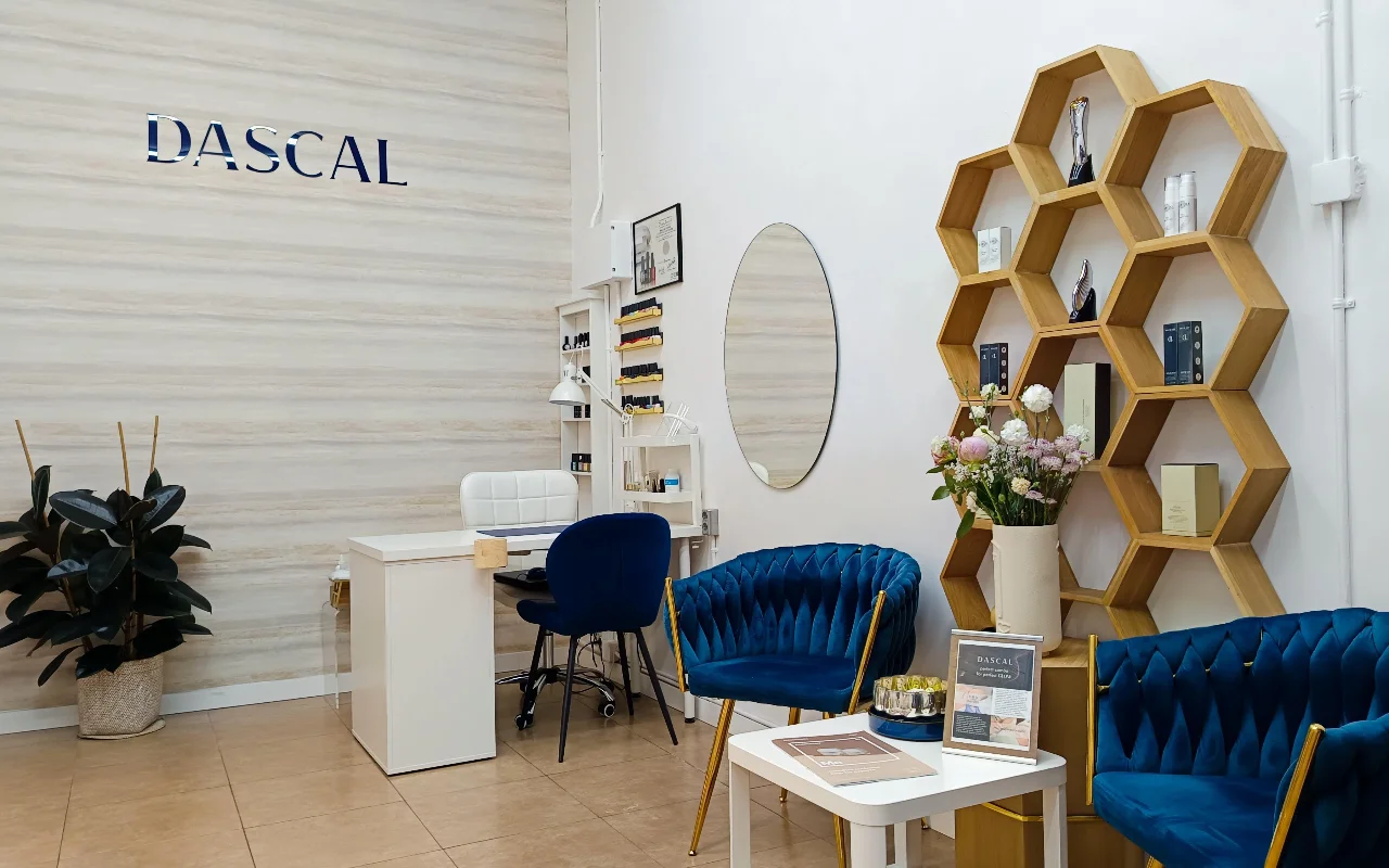 Een stijlvol salon-interieur met diepblauwe fluwelen stoelen en een bijzonder zeshoekig houten wandrek. In de hoek staat een witte manicuretafel tegen een gestructureerde, lichtgekleurde muur met het woord "DASCAL" in blauwe letters.
