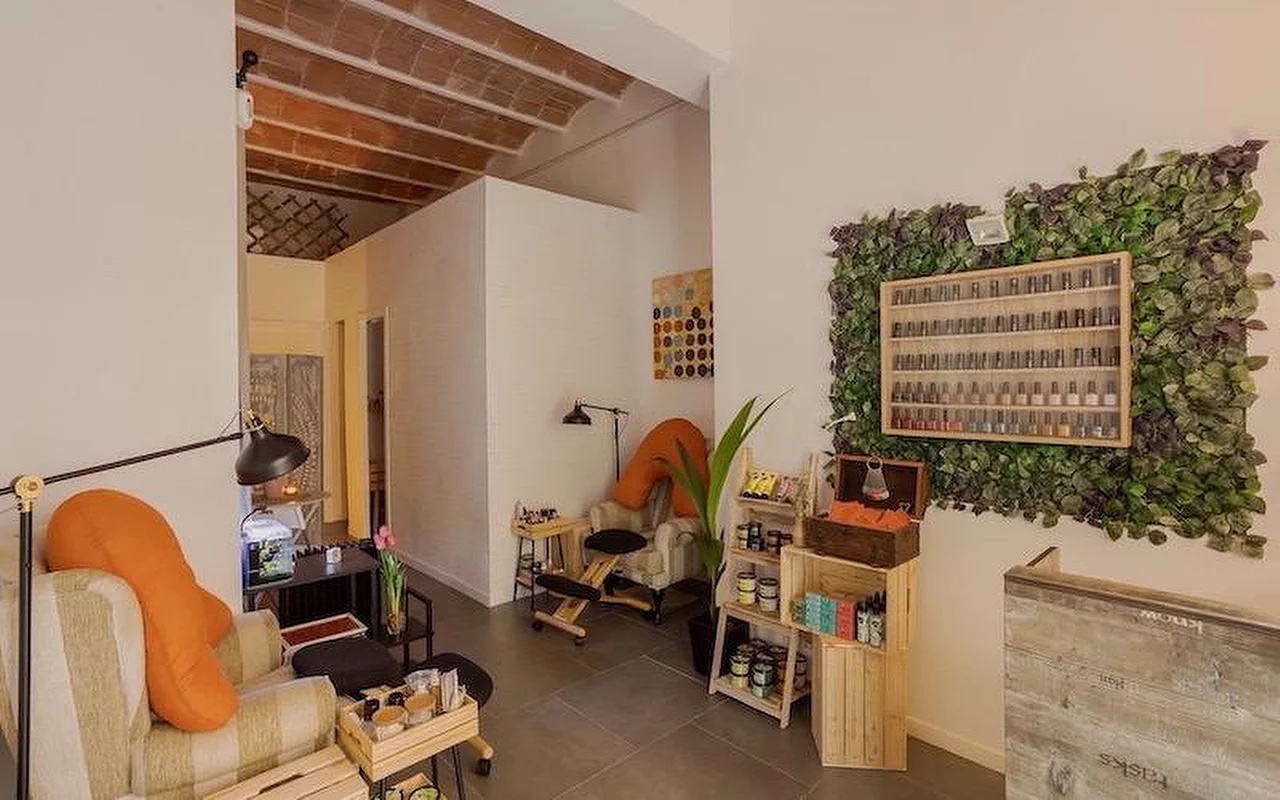 Een gezellige nagelsalon in bohemien-stijl met zachte fauteuils en oranje kussens. Aan de muur hangt een grote verticale planteninstallatie (groene muur) rondom een houten vitrinekast gevuld met nagellakflesjes.