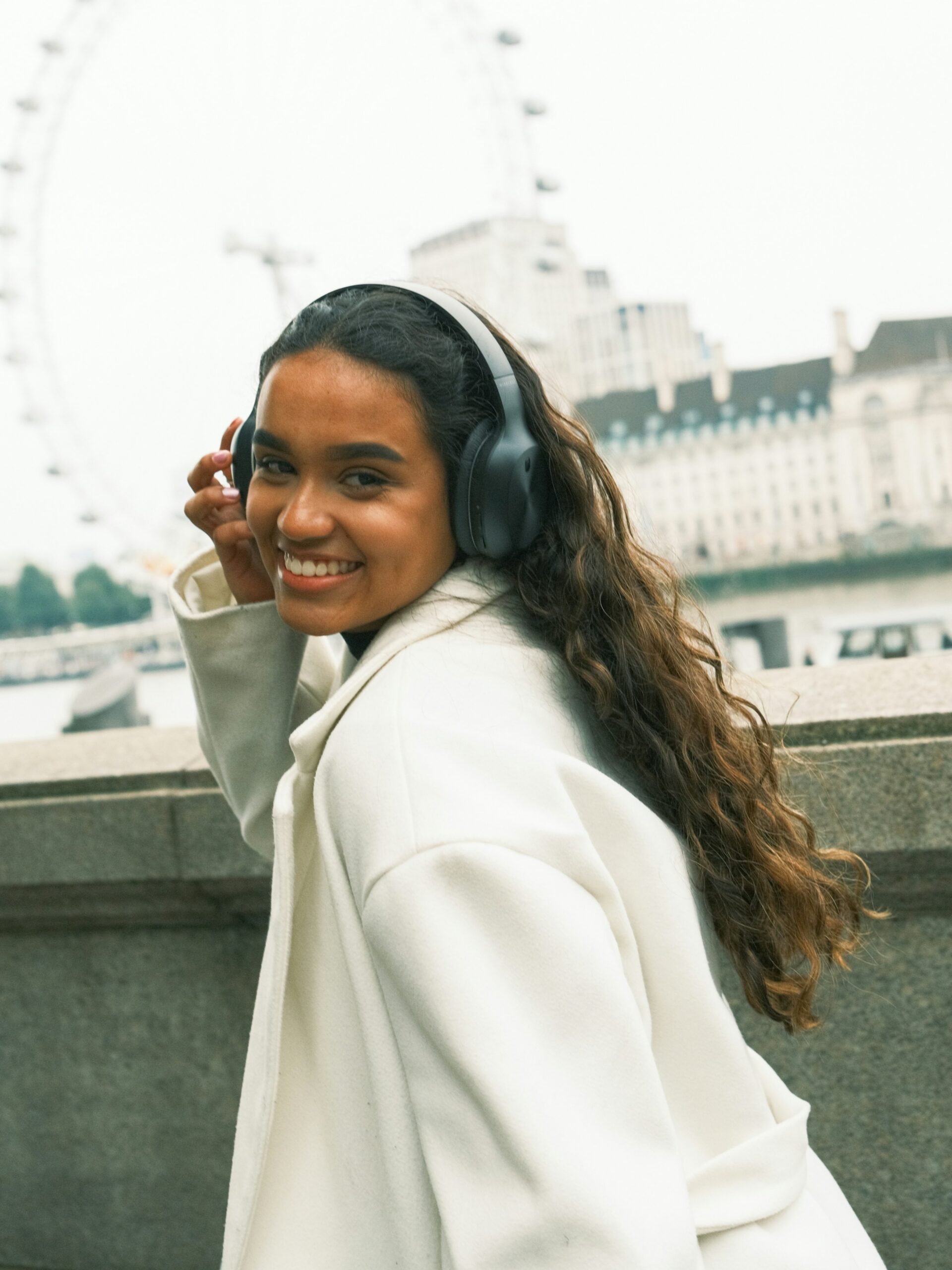 Een lachende vrouw met microblading op haar wenkbrauwen, met een zwarte over-ear koptelefoon en witte jas, wandelend langs de Theems in Londen