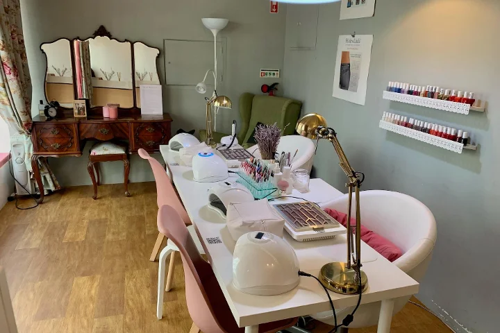 Het interieur van een gezellige beautysalon gespecialiseerd in manicures. Op de voorgrond een lange witte tafel met nagellampen, bureaulampen en gereedschap voor nageltechnici, geflankeerd door roze en witte stoelen. Op de achtergrond een vintage houten kaptafel met een drieluikspiegel, een groene fauteuil en witte planken aan de muur met rijen kleurrijke nagellakflesjes.