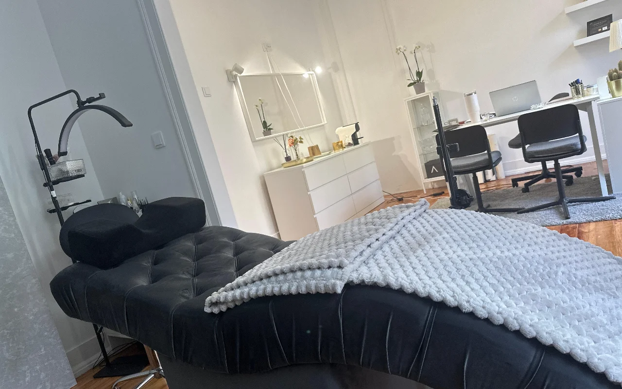 Een moderne wimperbehandelkamer vanuit een laag camerastandpunt. Op de voorgrond een luxe, zwart gecapitonneerde beautybank met een getextureerde grijze deken en een ergonomisch nekkussen onder een gebogen ringlamp. Op de achtergrond een minimalistische witte kantooropstelling met een bureau, twee zwarte stoelen, een laptop, een witte ladekast en een grote spiegel.