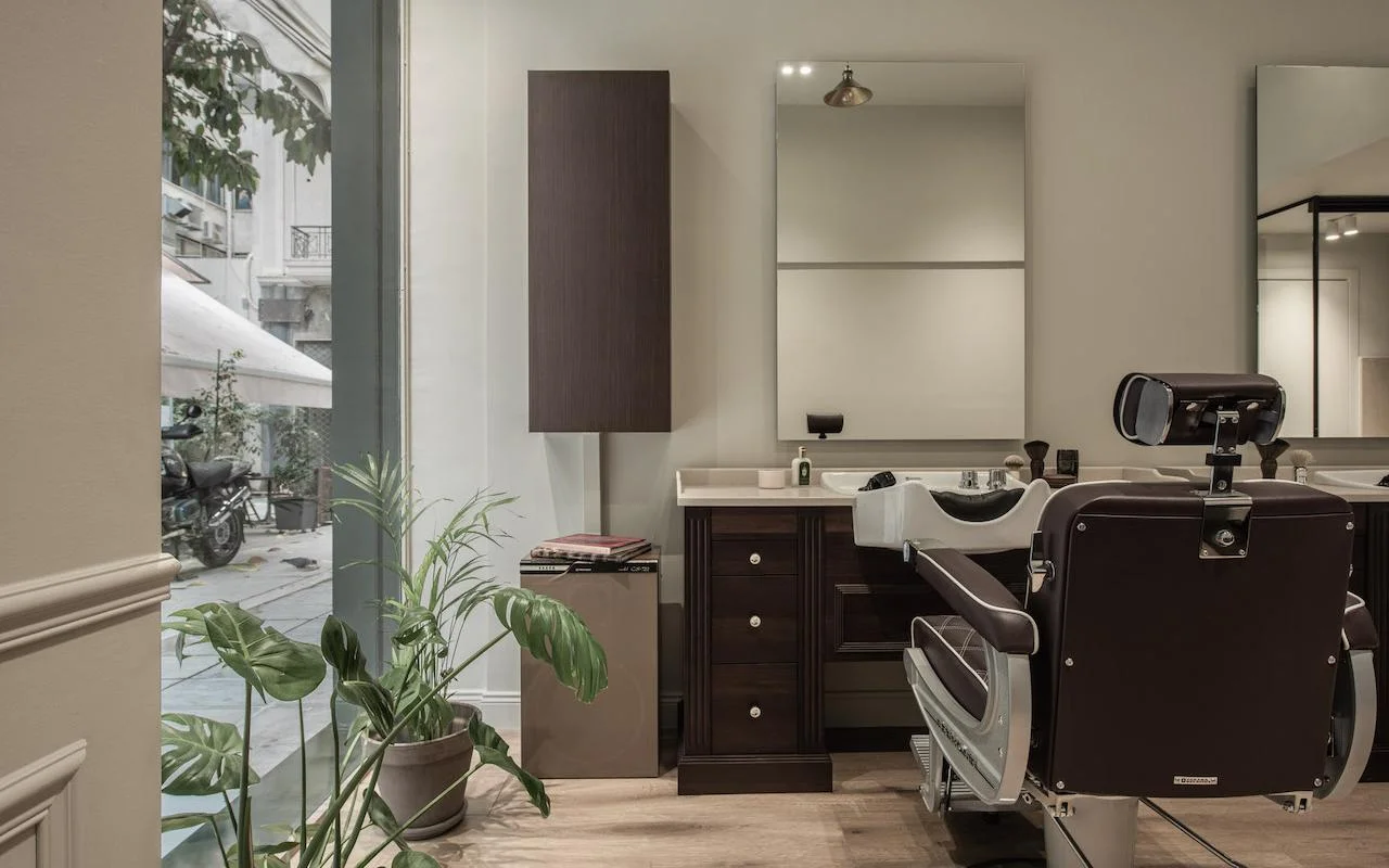 Een verfijnde barbershop met een donkerbruine leren kappersstoel, notenhouten kasten, een groot raam met uitzicht op een straat in Kolonaki, en een monstera-plant die groen toevoegt aan de minimalistische ruimte.
