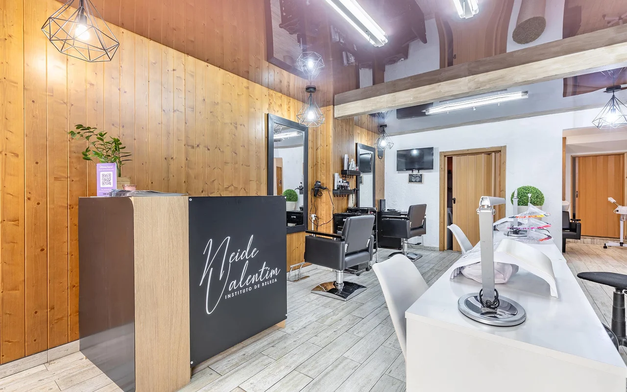 Een eigentijdse beautysalon met een receptiebalie links, gebranded met 'Neide Valentim Instituto de Beleza'. De muren zijn afgewerkt met warme verticale houten panelen en grote spiegels. Zwarte kappersstoelen staan voor de spiegels, terwijl een witte manicurestation met witte stoelen aan de rechterkant langs de grijze tegelvloer loopt.