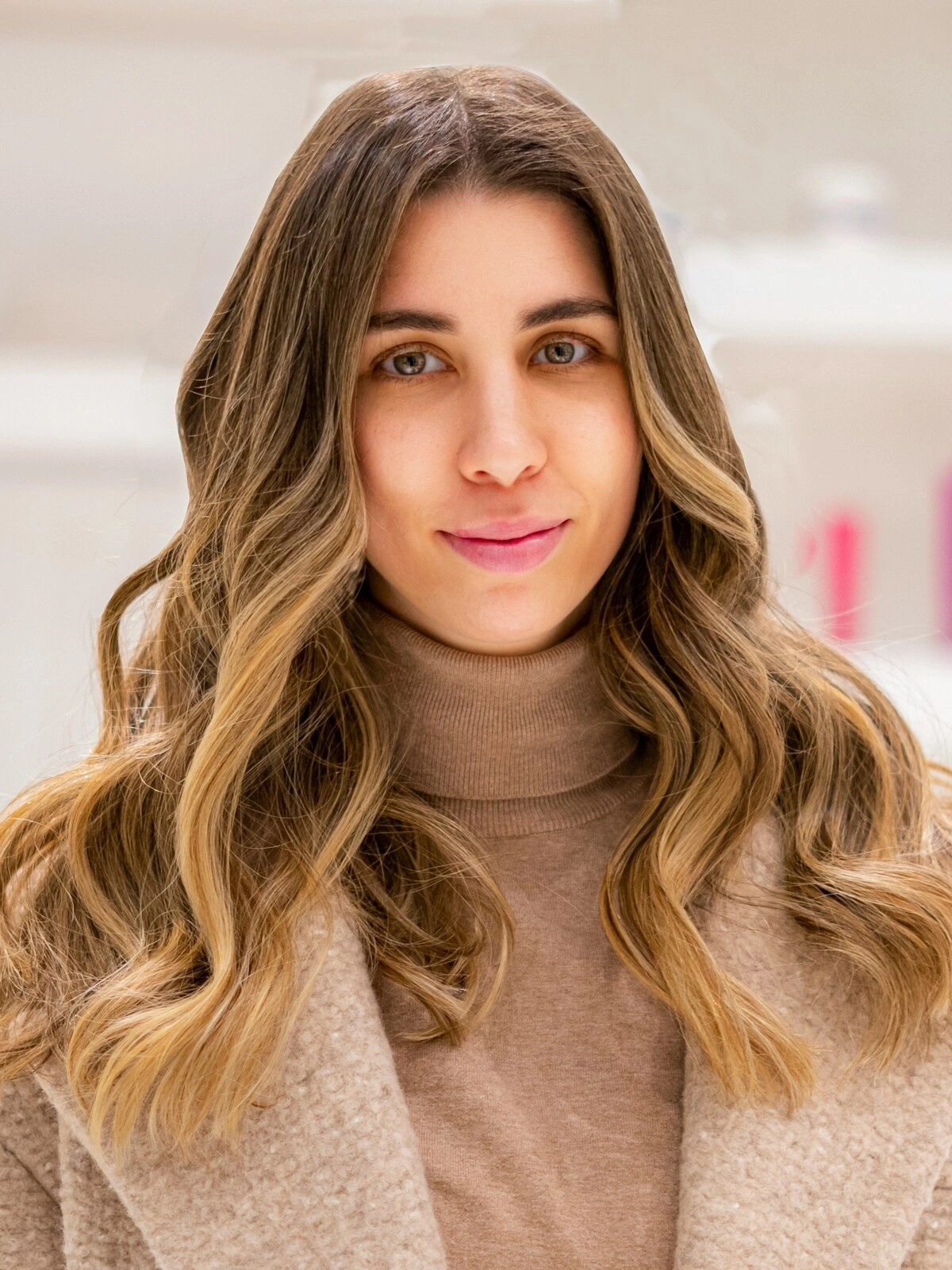 Een vrouw in een coltrui en een wollen jas in kamelkleur, met zachte, golvende balayage waarbij het bruin overloopt in karameltinten
