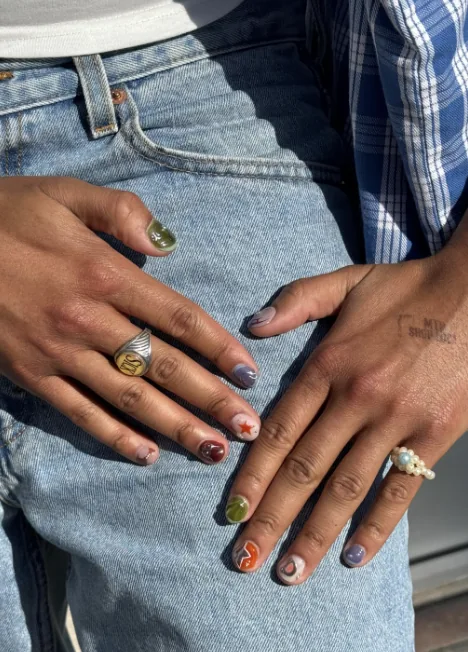 Korte mismatch-manicure waarbij elke nagel een andere kleur en ontwerp heeft, met een rode ster en een oranje swirl, rustend op een spijkerbroek.