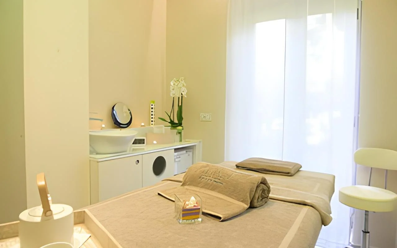 Een rustige behandelkamer bij Alice Beauty Clinique met een bed in zacht taupe linnen, witte orchideeën op het werkblad, kaarsen bij de wasbak en vitrage die het daglicht door een groot raam filtert.