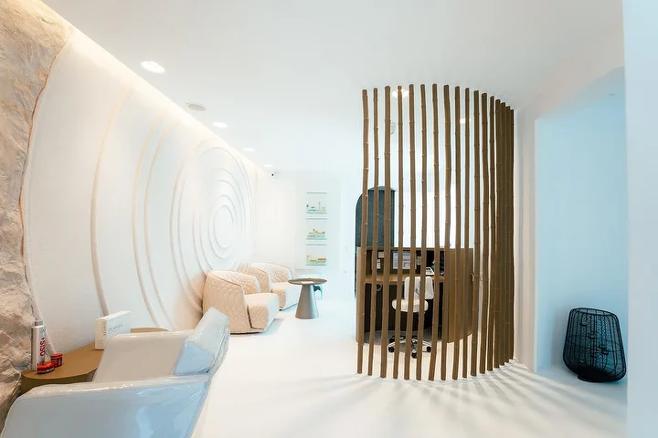 Een rustig, grotachtig spa-interieur met gebogen witte muren, waarvan er een is uitgesneden in concentrische rimpels, zachte indirecte verlichting, gewatteerde crèmekleurige fauteuils en een gebogen houten lamellenscherm dat een rustige behandelhoek afschermt.