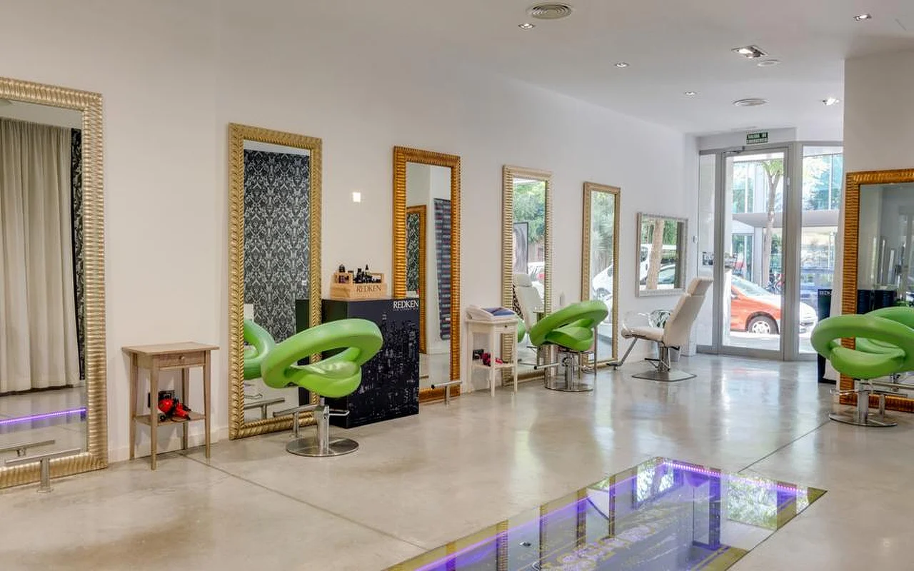 Een lichte, ruime kapsalon met gepolijste betonvloeren, hoge spiegels met gouden lijsten langs een witte muur, limoengroene gevormde kappersstoelen en een glazen vloergedeelte met paars licht, met de straat zichtbaar door de glazen deuren achterin.