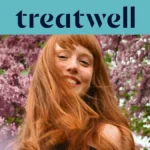 Close-up portret van een glimlachende jonge roodharige vrouw met een pony, wiens haar in de wind waait voor een achtergrond van roze seringentakken. Een lichtblauwe banner bovenaan toont het "Treatwell"-logo in het donkerblauw.