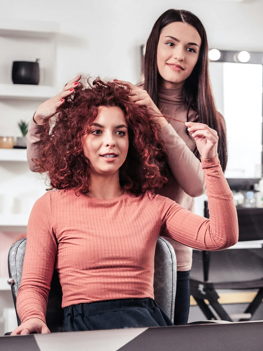 Een klant met geverfd rood krullend haar zit bij de kapper terwijl haar stylist de behandeling bespreekt