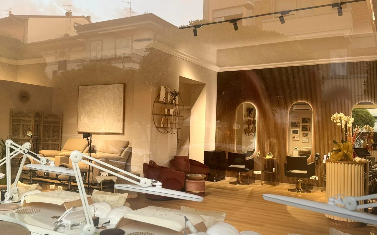 Het interieur van de salon van Lilian Almanza, gezien door de etalage tijdens het gouden uur. Aan de linkerkant staan nagelstations met verstelbare lampen, rechts zie je warme houten lambrisering, boogvormige spiegels met verlichte lijsten en fluwelen fauteuils.