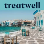 De boulevard van Klein Venetië in Mykonos-stad: witgekalkte huizen met kleurige houten balkons aan het water, daarachter de turkooizen Egeïsche Zee en op de voorgrond witte en petrolblauwe cafétafels op een geplaveid terras.