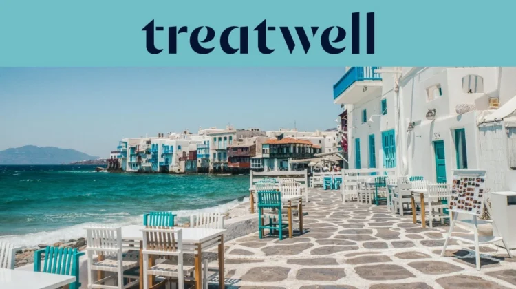 De boulevard van Klein Venetië in Mykonos-stad: witgekalkte huizen met kleurige houten balkons aan het water, daarachter de turkooizen Egeïsche Zee en op de voorgrond witte en petrolblauwe cafétafels op een geplaveid terras.