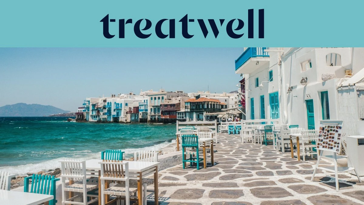 De boulevard van Klein Venetië in Mykonos-stad: witgekalkte huizen met kleurige houten balkons aan het water, daarachter de turkooizen Egeïsche Zee en op de voorgrond witte en petrolblauwe cafétafels op een geplaveid terras.