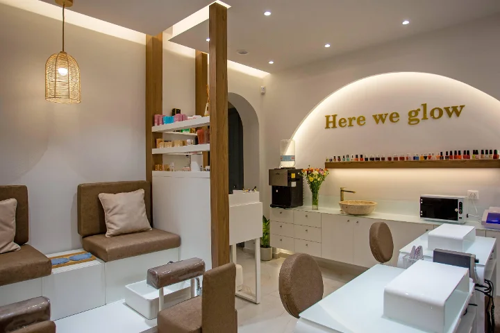 Een warme, zacht verlichte nagelsalon met een gevlochten rotan hanglamp, gestoffeerde bruine pedicurebanken, witte manicuretafels, een schap met kleurige lakjes en een gouden 'Here we glow'-bord op een achtergrond verlichte boog.