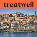 De Ribeira-kade in Porto, met rijen kleurrijke huizen en terracotta daken die vanaf de rivier de Douro tegen de heuvel op lopen, en de Clérigos-toren aan de horizon onder een strakblauwe lucht.
