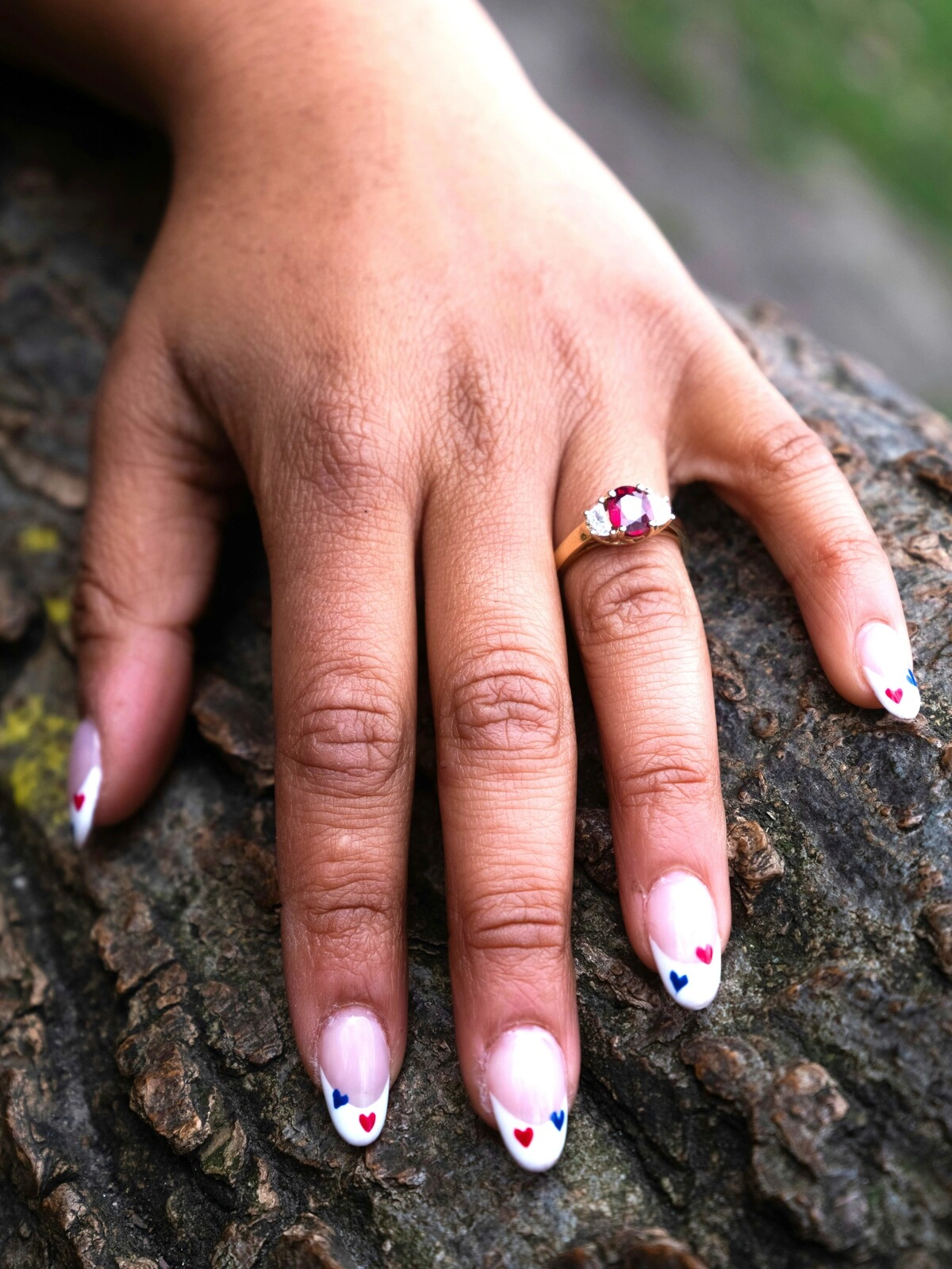 De hand van een vrouw rust op een tak en toont een French manicure op ovale nagels, versierd met kleine rode en blauwe hartjes