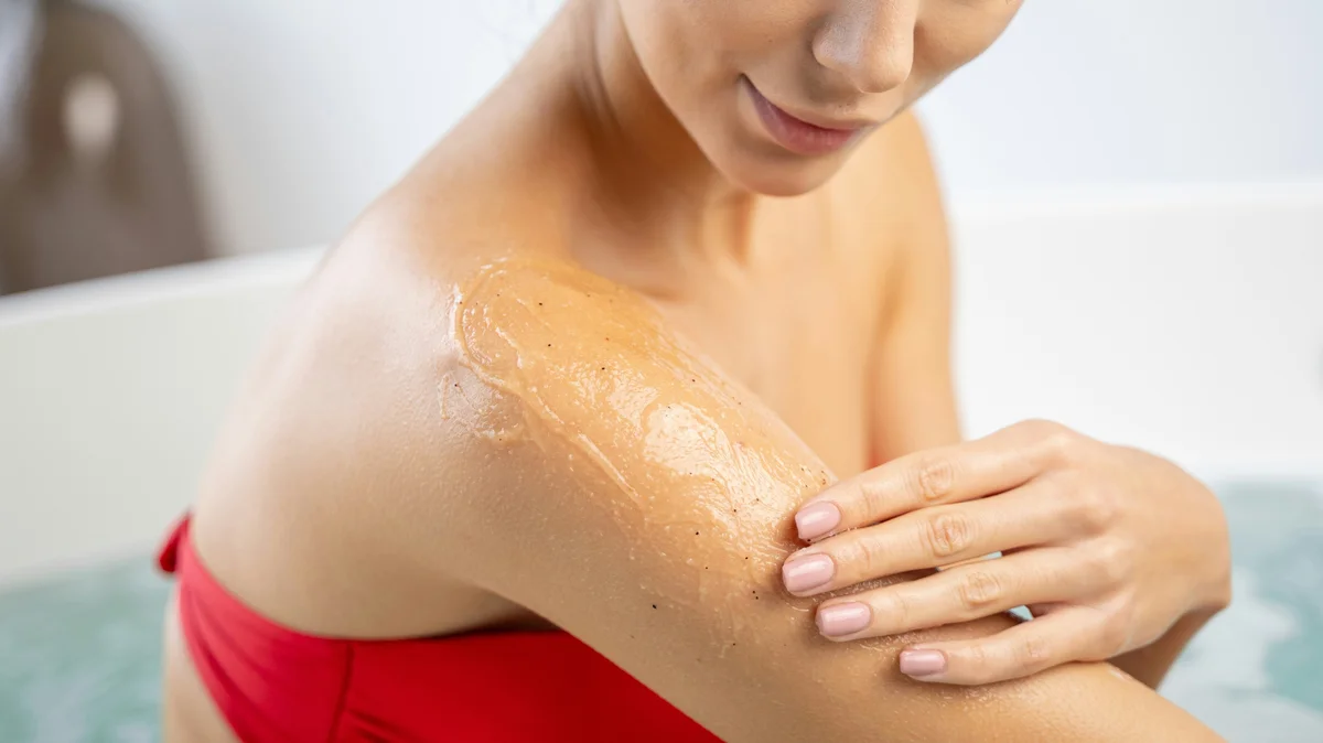 Close-up van een vrouw in een rode bikini die een scrub aanbrengt op haar bovenarm in een lichte, opgeruimde badkamer