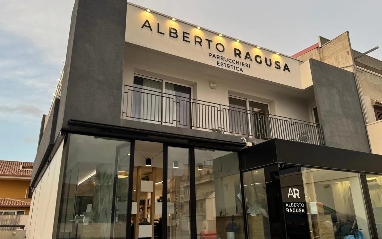 De buitenkant van Alberto Ragusa in de schemering, een salon van twee verdiepingen met een groot verlicht bord met ‘Alberto Ragusa Parrucchieri Estetica’ boven vloer-tot-plafondramen, met warm licht dat van binnenuit schijnt.