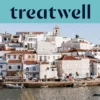 Een witgekalkt vissersdorp aan de Algarve met huizen met terracotta daken die tegen een heuvel omhoogklimmen boven een rustig water, kleine vissersboten afgemeerd langs de kade, palmbomen en een kerktoren onder een strakblauwe lucht.