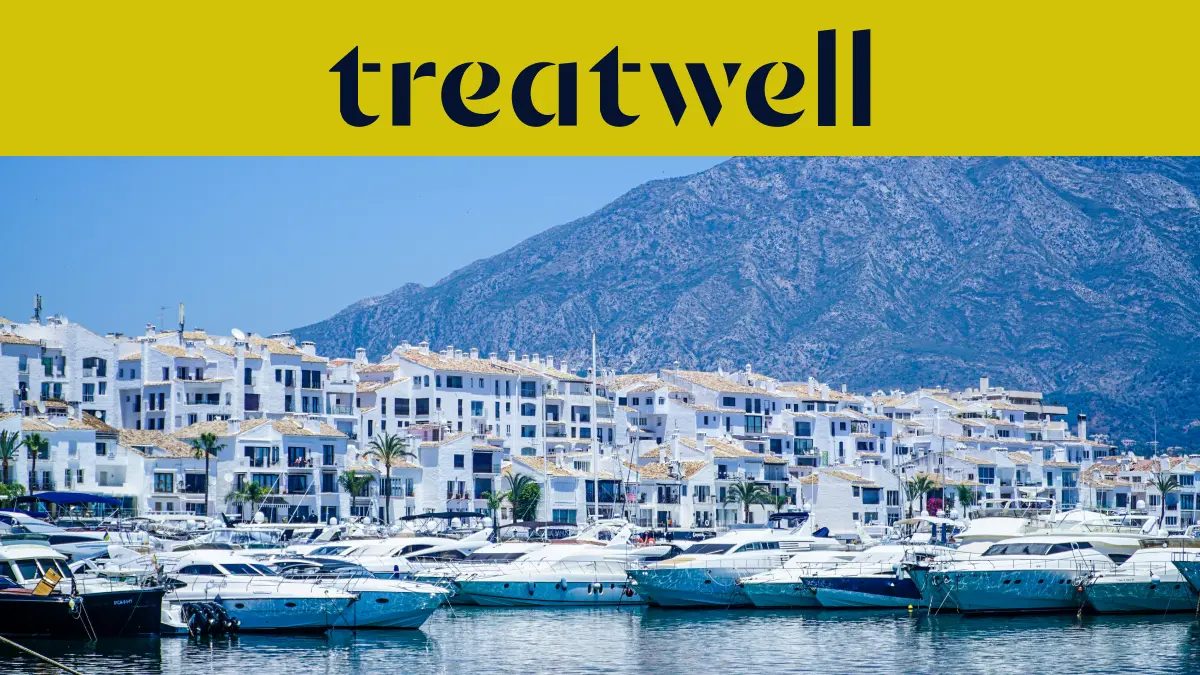 De jachthaven van Puerto Banús in Marbella, omzoomd door witte appartementsgebouwen en afgemeerde jachten met bergen op de achtergrond, onder een geel Treatwell-spandoek.