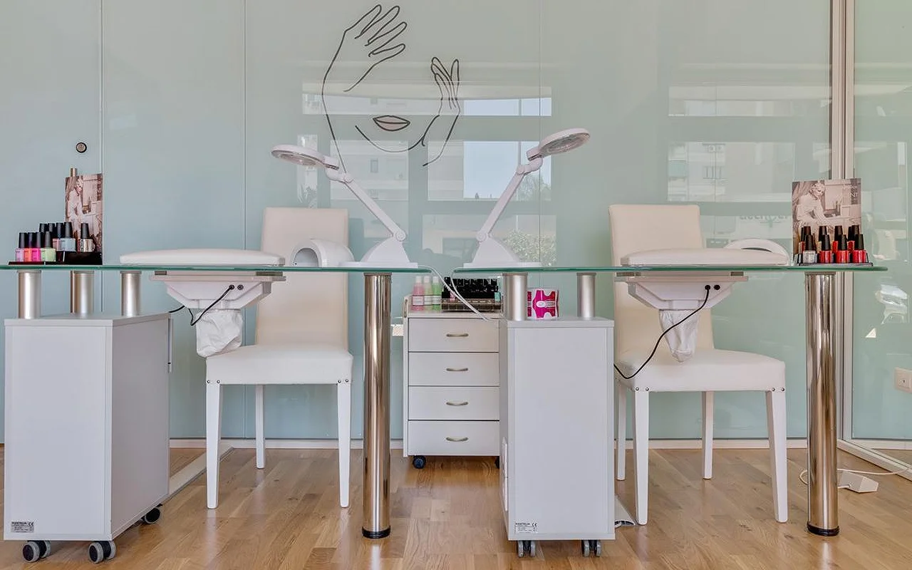 Een nagelstudio bij Perfetta Urban Beauty, met twee manicuretafels met glazen blad en witte stoelen, werklampen, kleurige lakdisplays en een mintgroene glaswand met de lijntekening van een gezicht en twee handen.