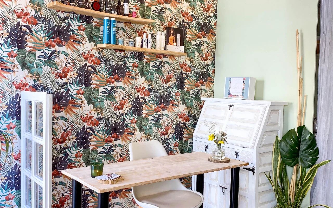 Een stylingplek bij Shangara Lora Organics in Marbella, met opvallend tropisch behang, een houten tafel, een oud wit secretaire en biologische haarproducten op zwevende planken.