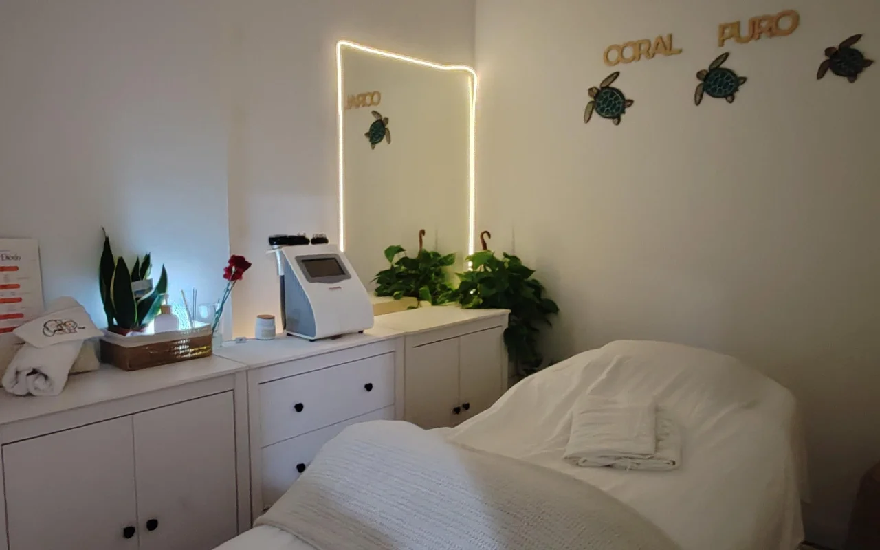 Een kleine, rustige behandelkamer met witte kasten en een bed opgemaakt met witte handdoeken. Een verlichte spiegel en een behandelapparaat staan op de toonbank tussen groene planten, onder gouden „Coral Puro'-belettering en decoratieve schildpad-muurkunst.